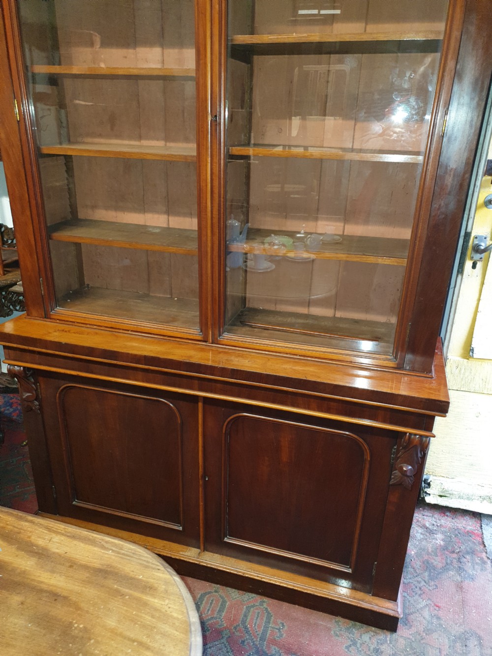 A Victorian mahogany library bookcase featuring upper double glazed doors, carved corner corbels, internal wooden shelving, and a lower two-door panelled cupboard base. View III