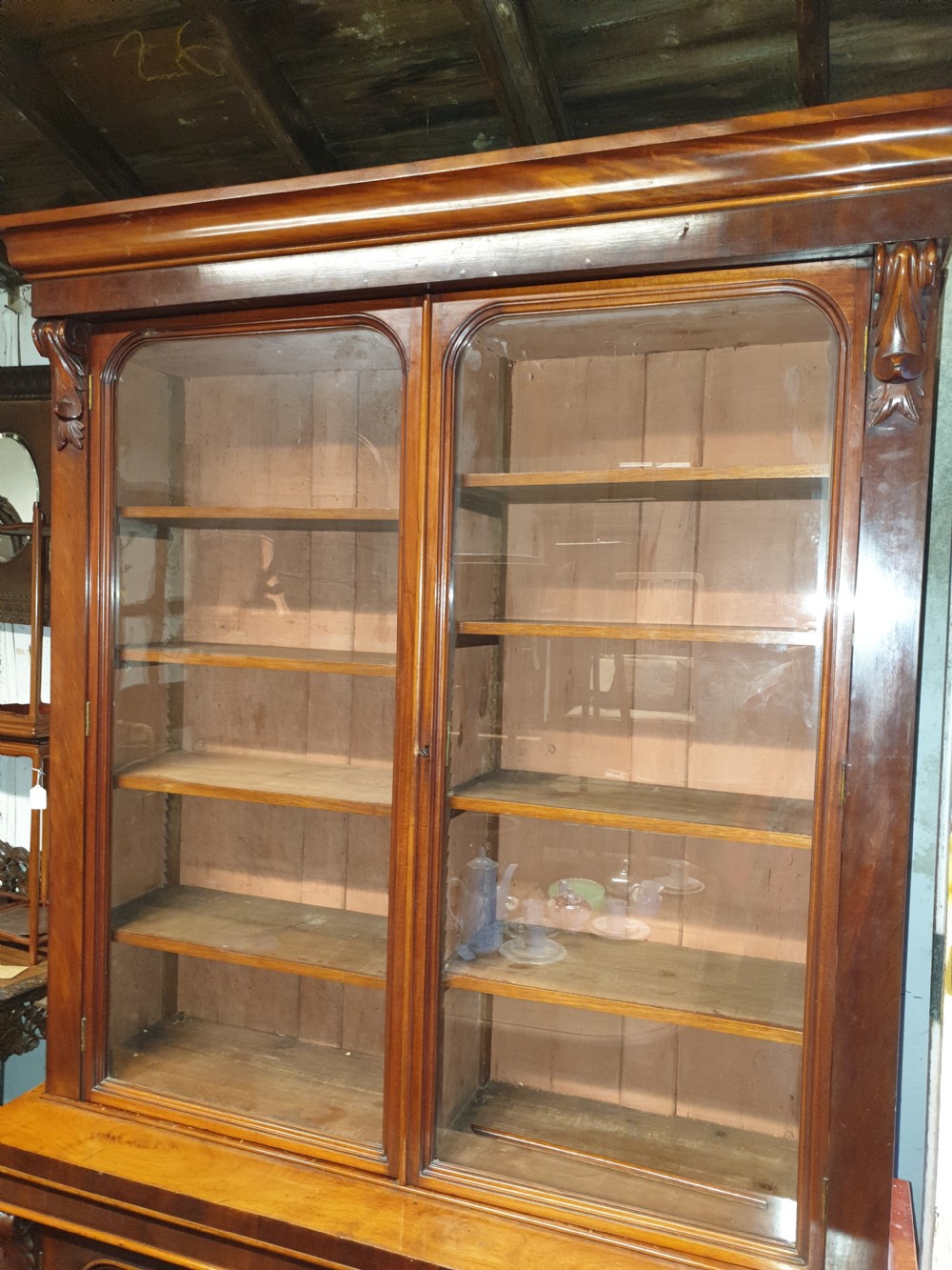 A Victorian mahogany library bookcase featuring upper double glazed doors, carved corner corbels, internal wooden shelving, and a lower two-door panelled cupboard base. View V