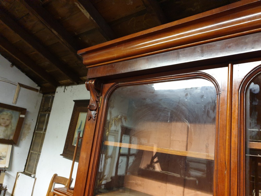 A Victorian mahogany library bookcase featuring upper double glazed doors, carved corner corbels, internal wooden shelving, and a lower two-door panelled cupboard base. View VI