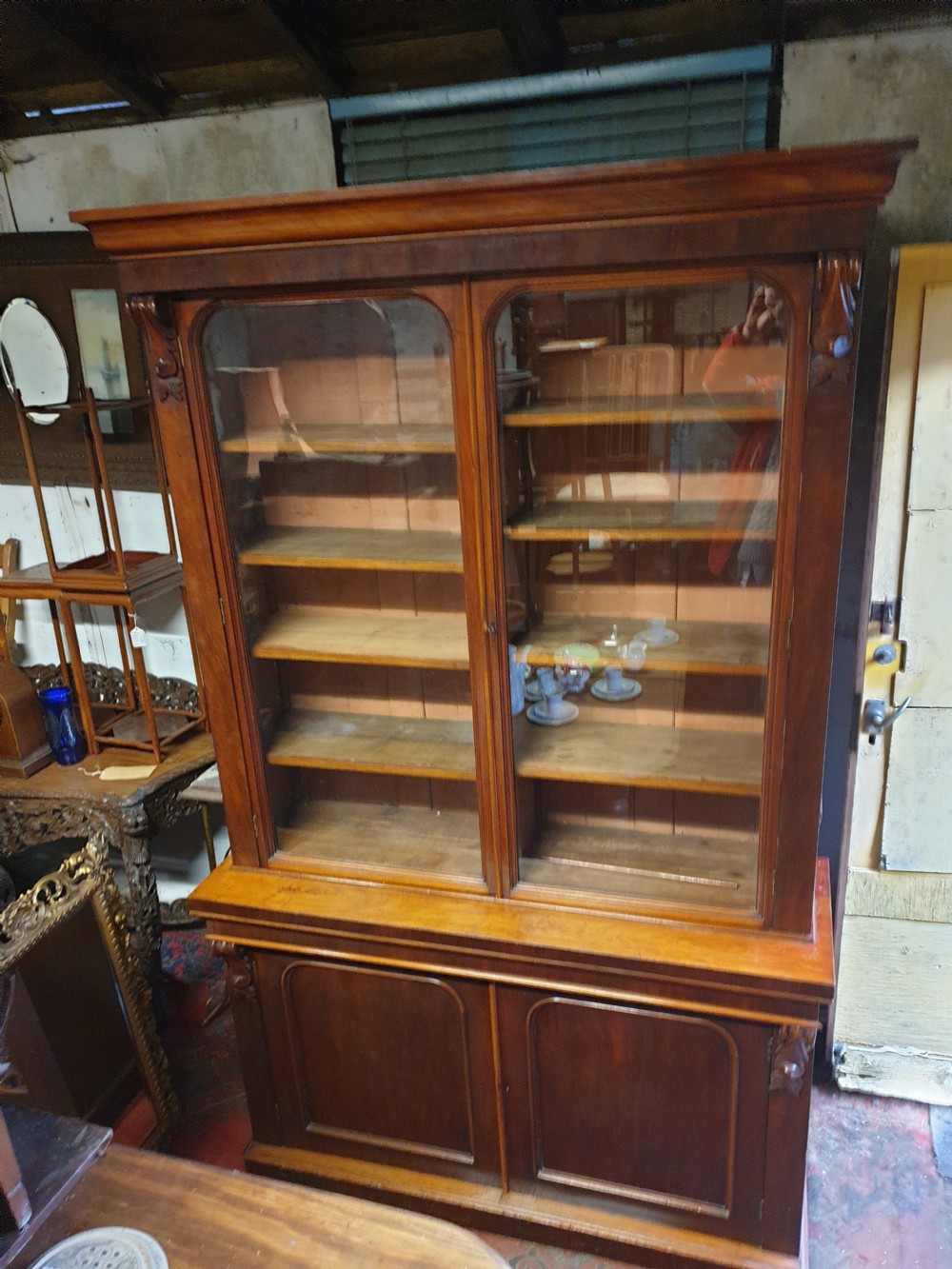 A Victorian mahogany library bookcase featuring upper double glazed doors, carved corner corbels, internal wooden shelving, and a lower two-door panelled cupboard base. View VII