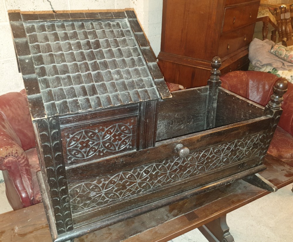 A dark carved oak Victorian rocking cradle featuring a tiled wooden hood, geometric floral carving, and turned finials.