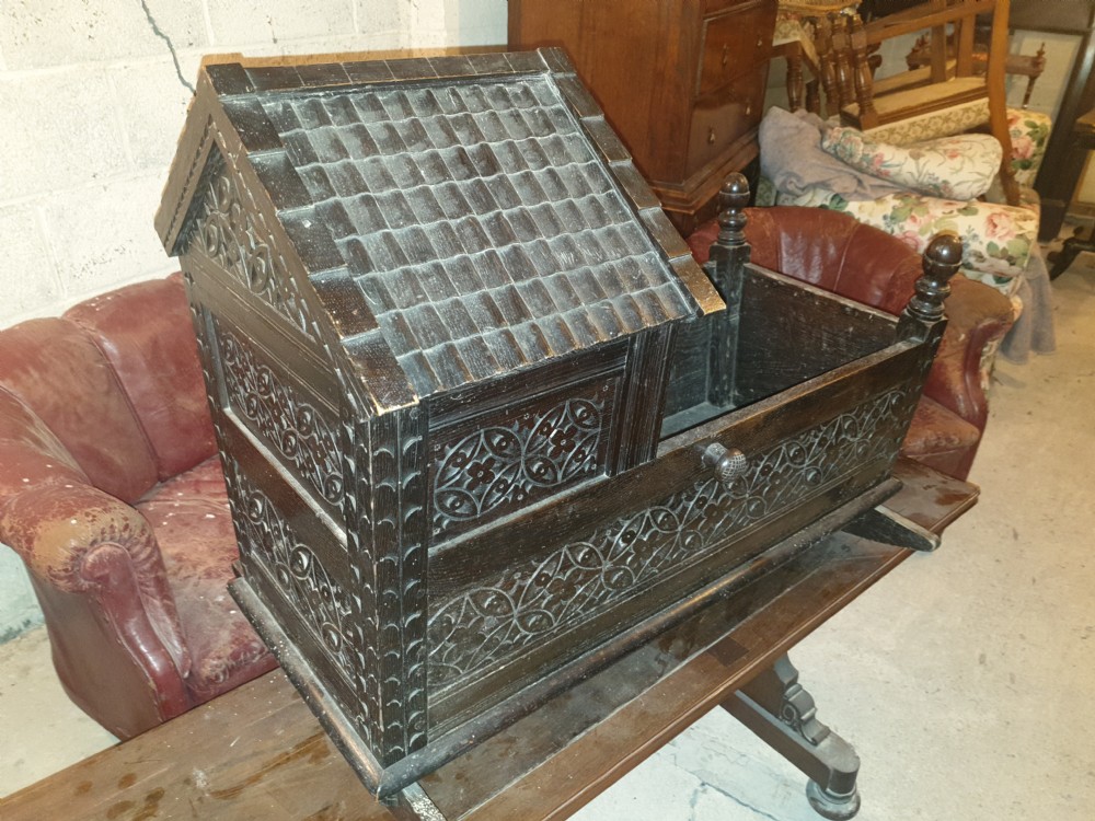 A dark carved oak Victorian rocking cradle featuring a tiled wooden hood, geometric floral carving, and turned finials. View VI