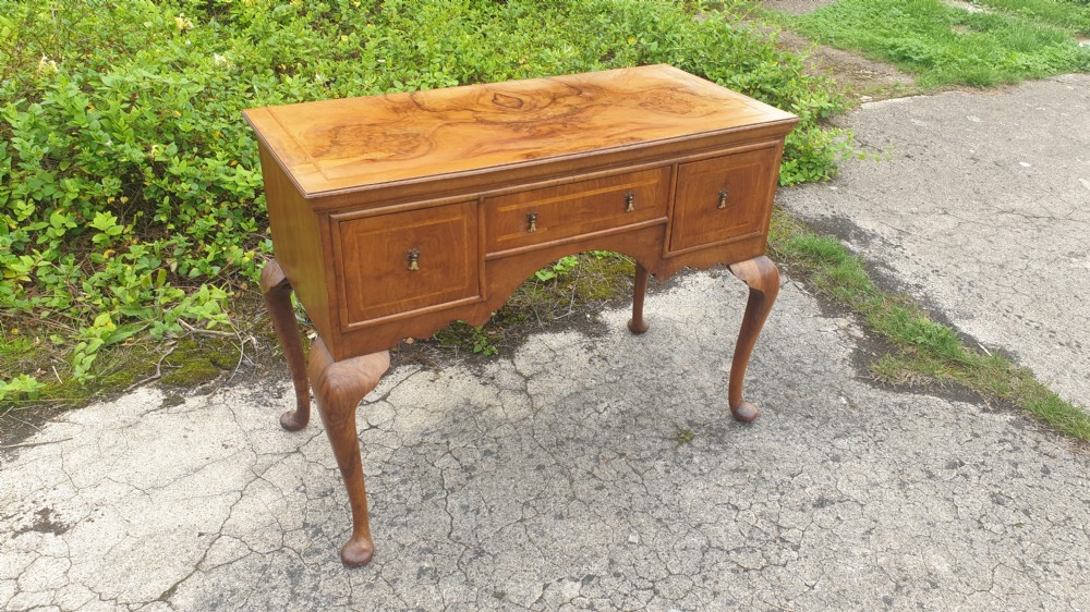 A Queen Anne style walnut side table featuring a figured veneer top, three drawers with brass handles, an arched apron, and four cabriole legs resting on pad feet.