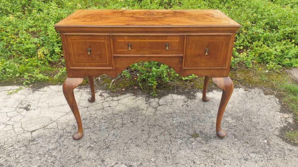 A Queen Anne style walnut side table featuring a figured veneer top, three drawers with brass handles, an arched apron, and four cabriole legs resting on pad feet. View VI