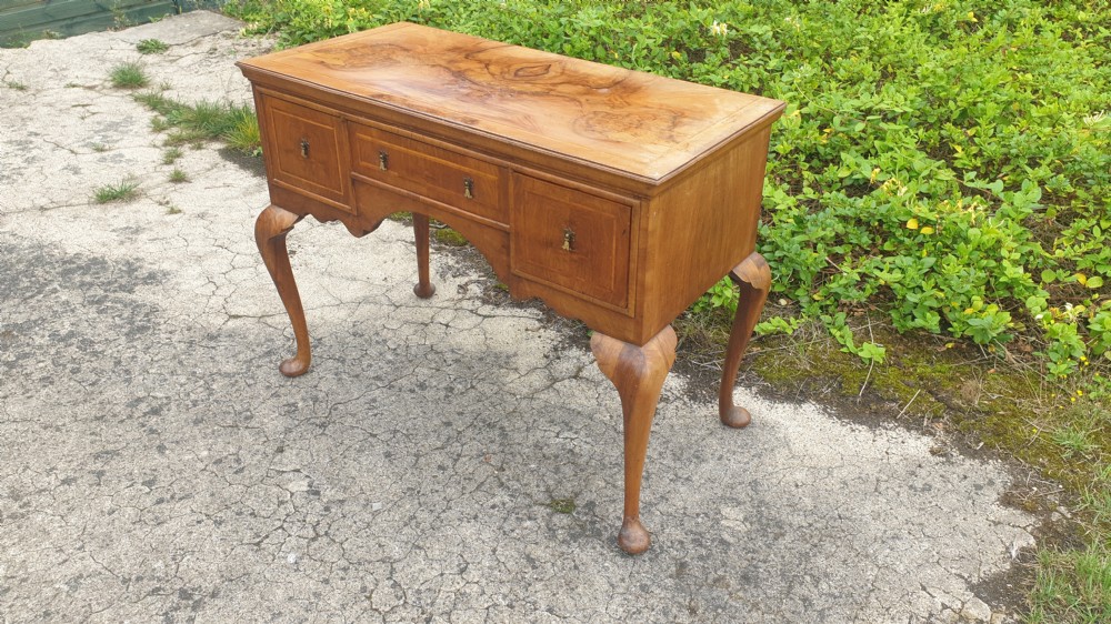 A Queen Anne style walnut side table featuring a figured veneer top, three drawers with brass handles, an arched apron, and four cabriole legs resting on pad feet. View VII