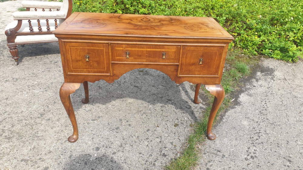 A Queen Anne style walnut side table featuring a figured veneer top, three drawers with brass handles, an arched apron, and four cabriole legs resting on pad feet. View VIII