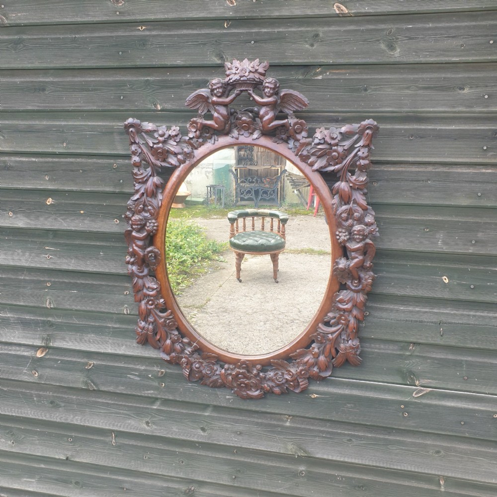 A late 19th-century oval wall mirror framed in dark, heavily carved walnut wood featuring motifs of cherubs, floral garlands, and a cresting basket.