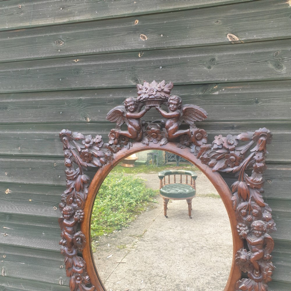 A late 19th-century oval wall mirror framed in dark, heavily carved walnut wood featuring motifs of cherubs, floral garlands, and a cresting basket. View II