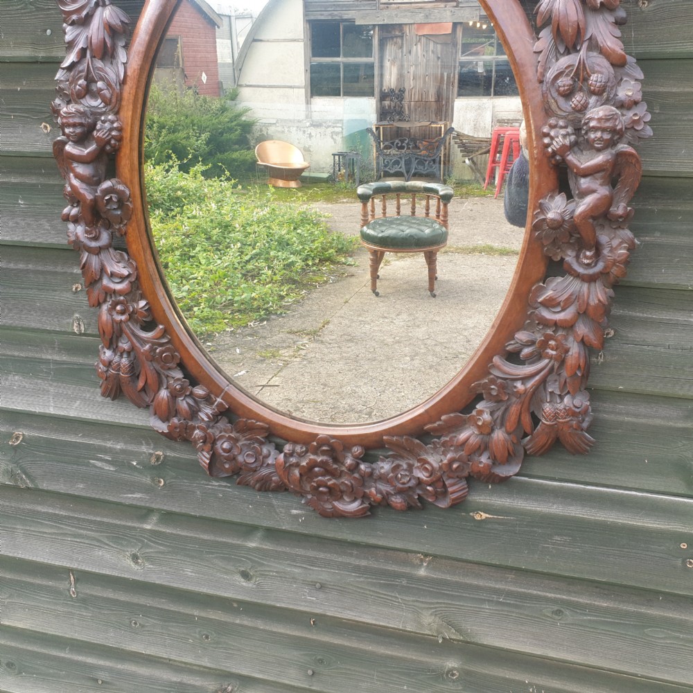 A late 19th-century oval wall mirror framed in dark, heavily carved walnut wood featuring motifs of cherubs, floral garlands, and a cresting basket. View III