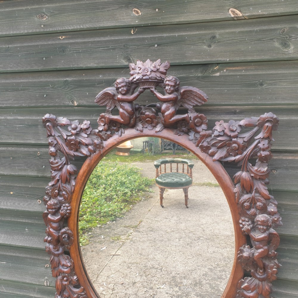A late 19th-century oval wall mirror framed in dark, heavily carved walnut wood featuring motifs of cherubs, floral garlands, and a cresting basket. View V