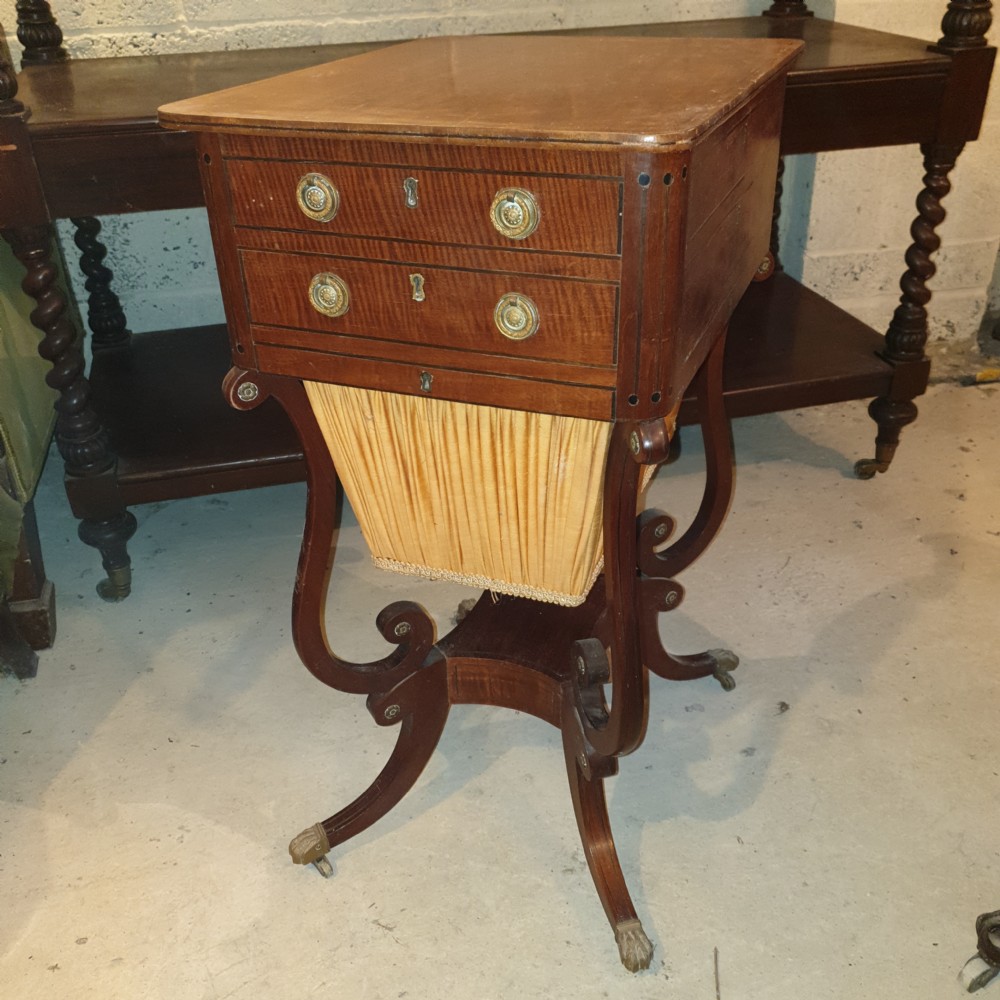 An antique Regency mahogany sewing table featuring inlaid drawers, a pleated fabric work basket, and swept legs with brass castors.