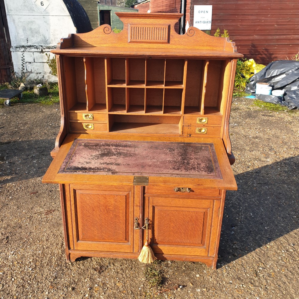 A late Victorian oak bureau cabinet featuring an open fall-front desk with an inset red leather writing surface, upper pigeonhole compartments, and lower double doors with brass handles. View IV