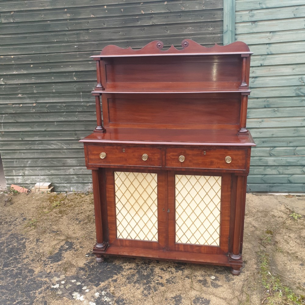 An antique Regency mahogany chiffonier cabinet featuring two raised shelves with turned supports, two drawers, and a double-door lower cabinet with brass lattice grilles and cream fabric backings.