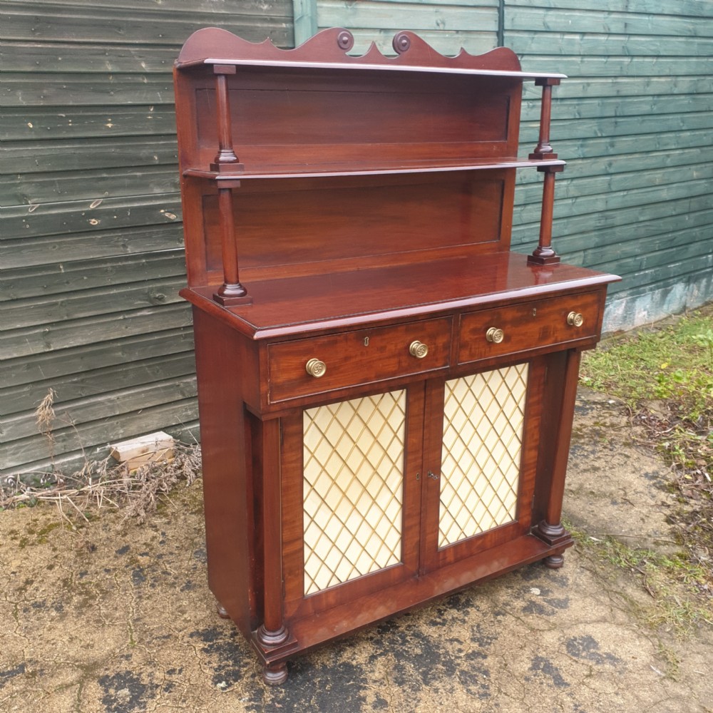 An antique Regency mahogany chiffonier cabinet featuring two raised shelves with turned supports, two drawers, and a double-door lower cabinet with brass lattice grilles and cream fabric backings. View V