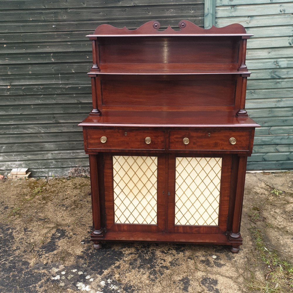 An antique Regency mahogany chiffonier cabinet featuring two raised shelves with turned supports, two drawers, and a double-door lower cabinet with brass lattice grilles and cream fabric backings. View VI
