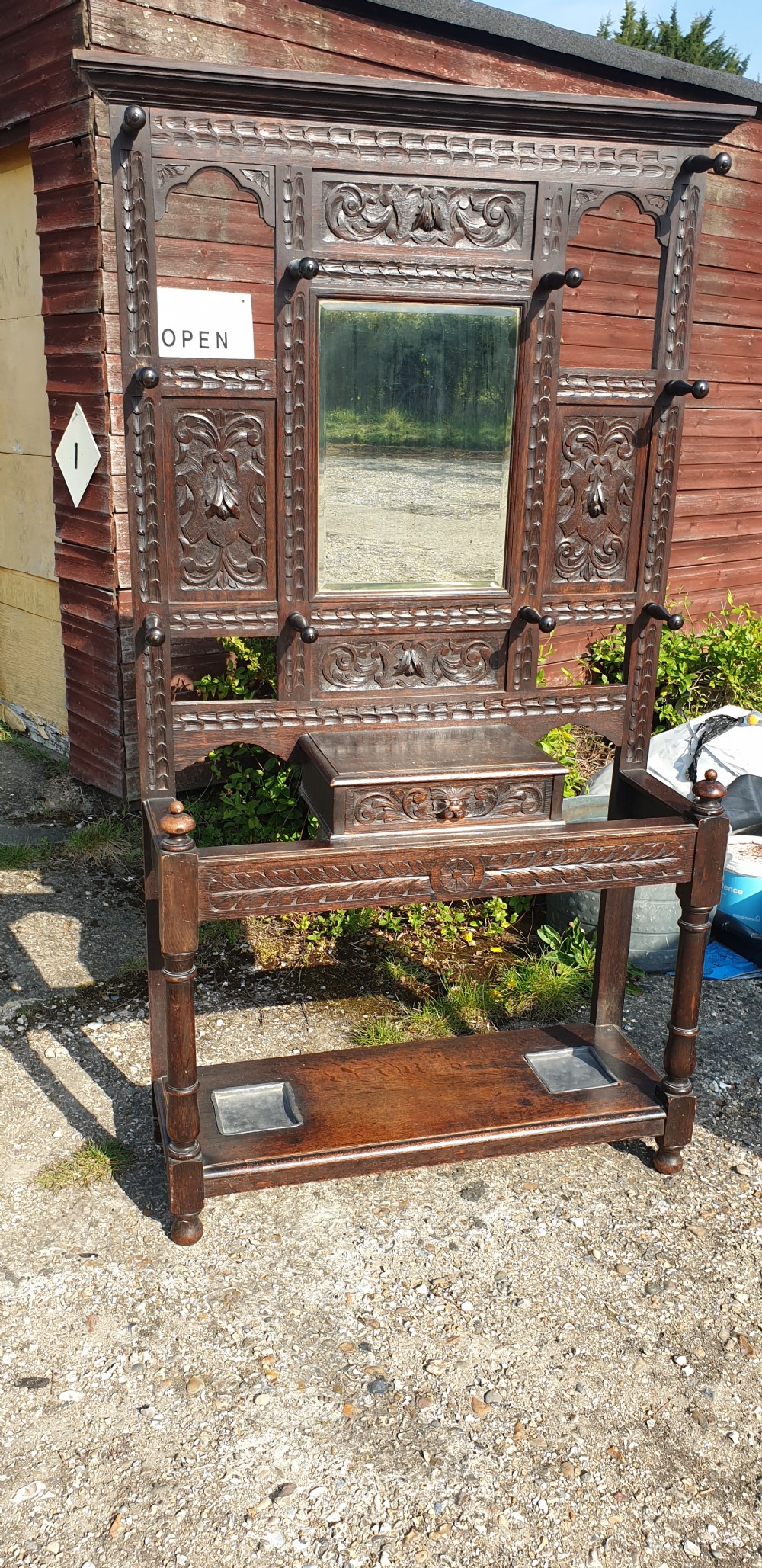 A late Victorian dark carved oak hallstand featuring a central mirror, carved Green Man motifs, coat pegs, a glove drawer, and integrated umbrella stands with metal drip trays. View II