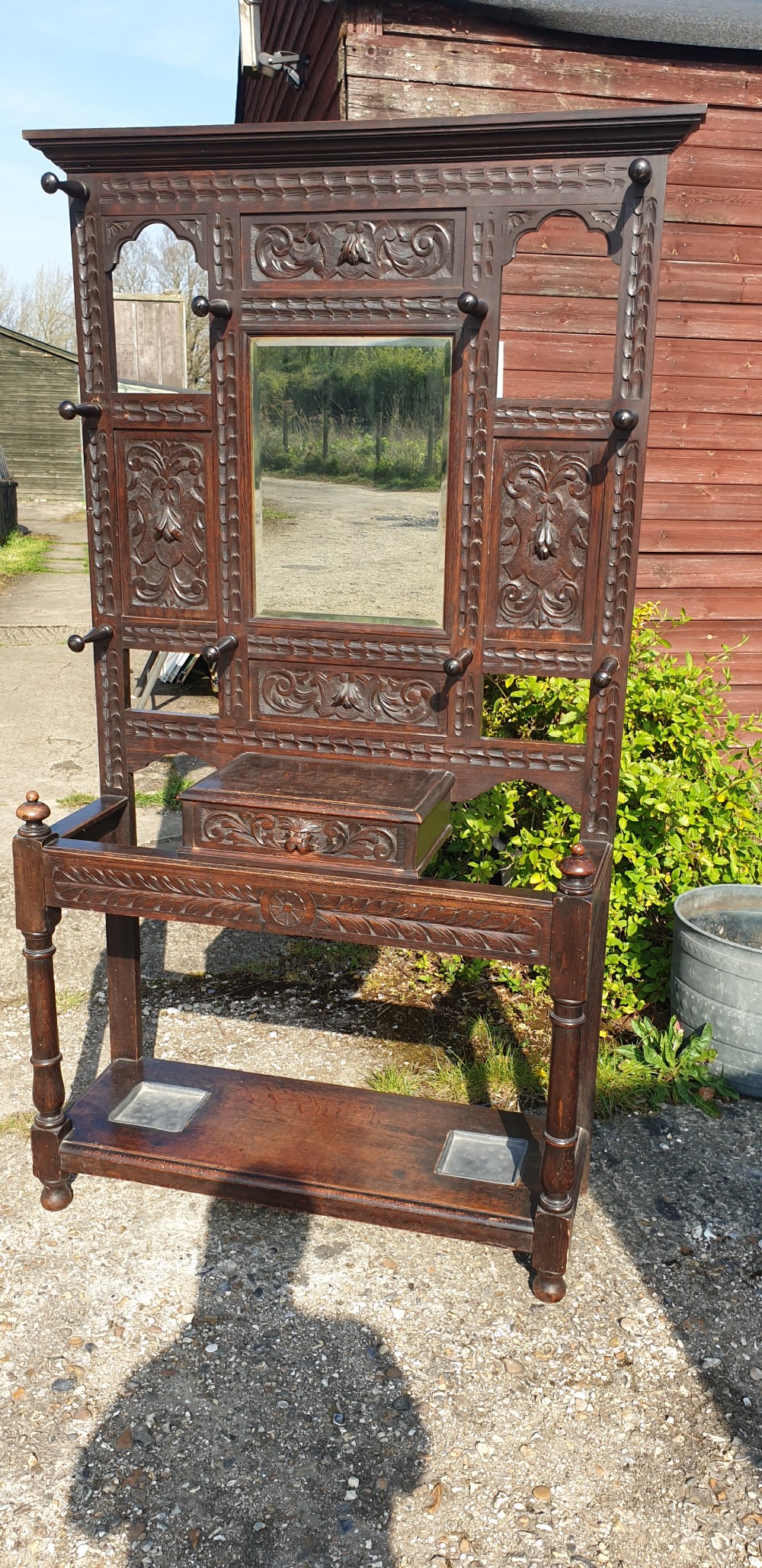 A late Victorian dark carved oak hallstand featuring a central mirror, carved Green Man motifs, coat pegs, a glove drawer, and integrated umbrella stands with metal drip trays. View III