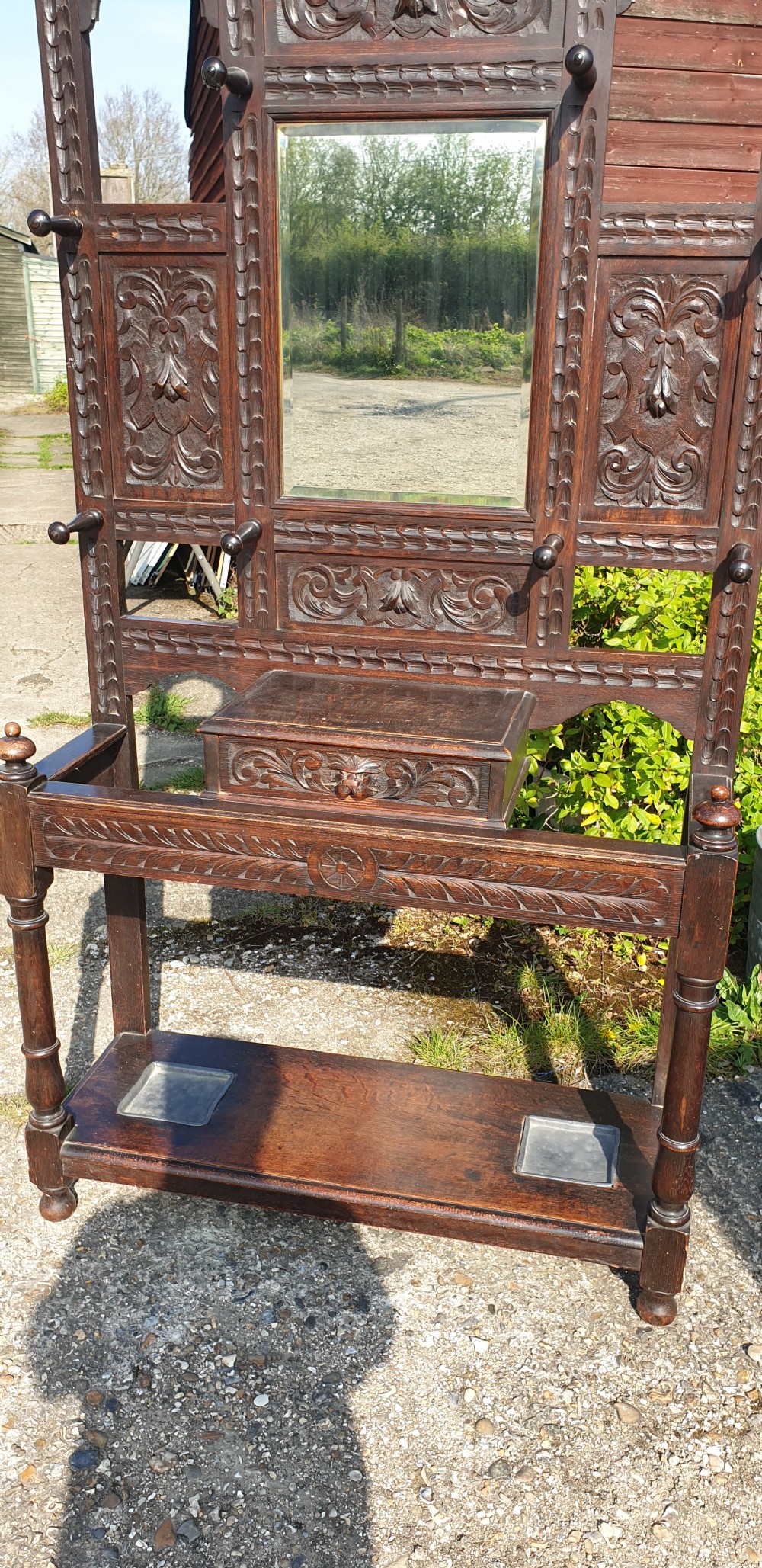A late Victorian dark carved oak hallstand featuring a central mirror, carved Green Man motifs, coat pegs, a glove drawer, and integrated umbrella stands with metal drip trays. View V