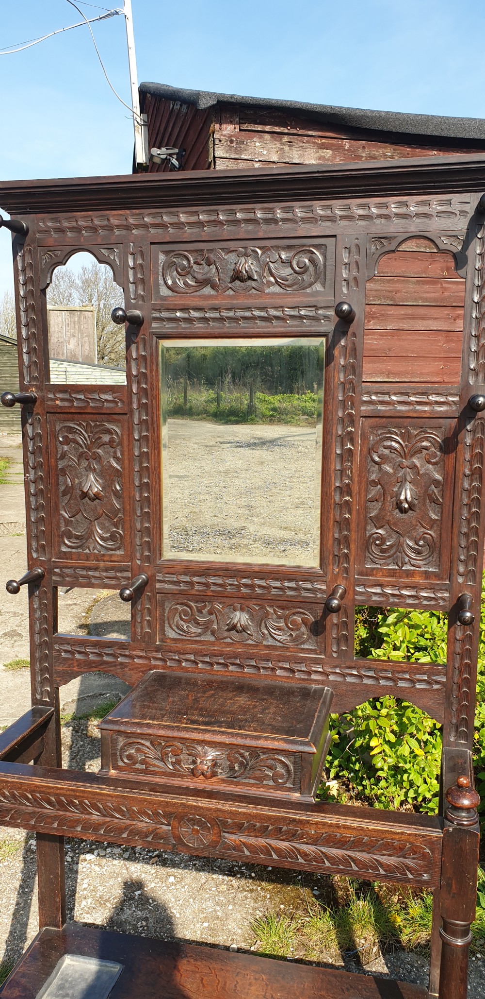 A late Victorian dark carved oak hallstand featuring a central mirror, carved Green Man motifs, coat pegs, a glove drawer, and integrated umbrella stands with metal drip trays. View VI
