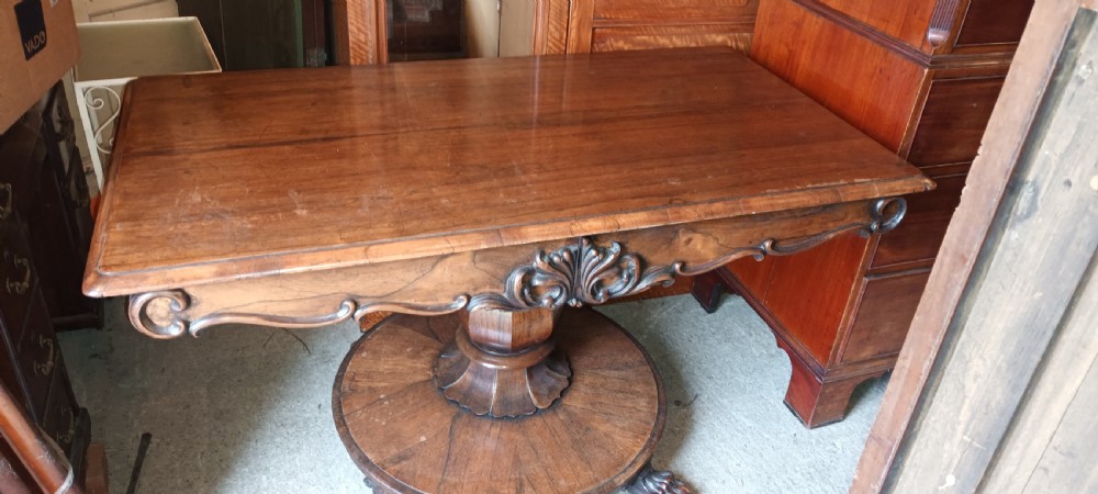 An early Victorian dark rosewood sofa table with a carved central apron motif, turned pedestal column, and circular platform base with scroll feet.