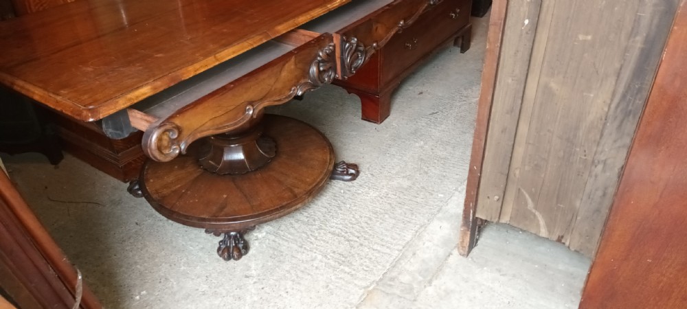 An early Victorian dark rosewood sofa table with a carved central apron motif, turned pedestal column, and circular platform base with scroll feet. View III