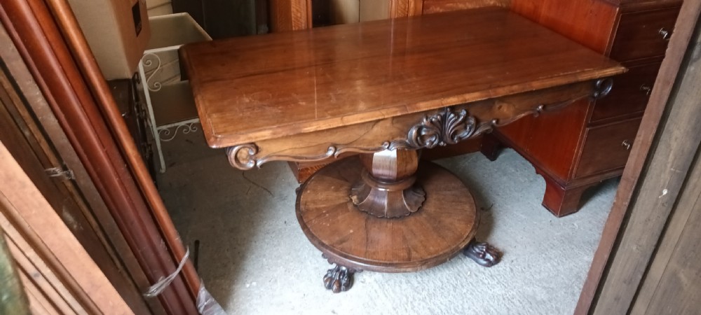 An early Victorian dark rosewood sofa table with a carved central apron motif, turned pedestal column, and circular platform base with scroll feet. View VI