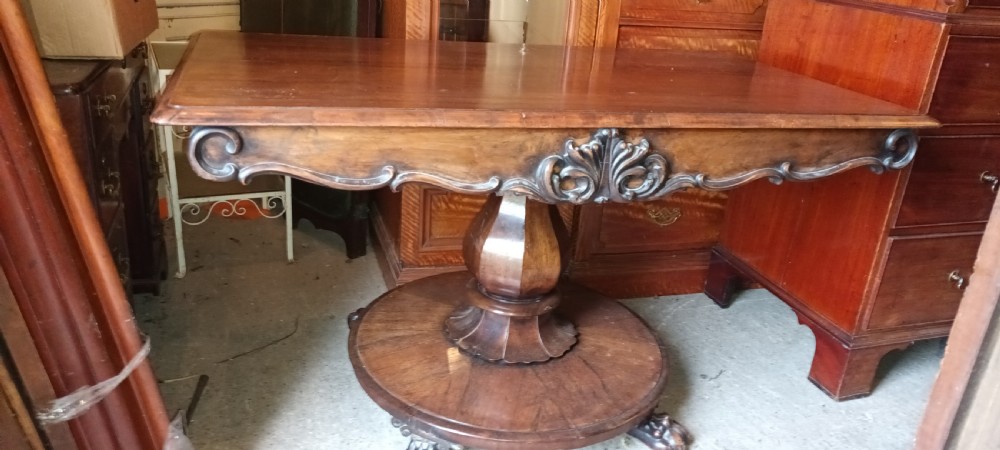 An early Victorian dark rosewood sofa table with a carved central apron motif, turned pedestal column, and circular platform base with scroll feet. View VII