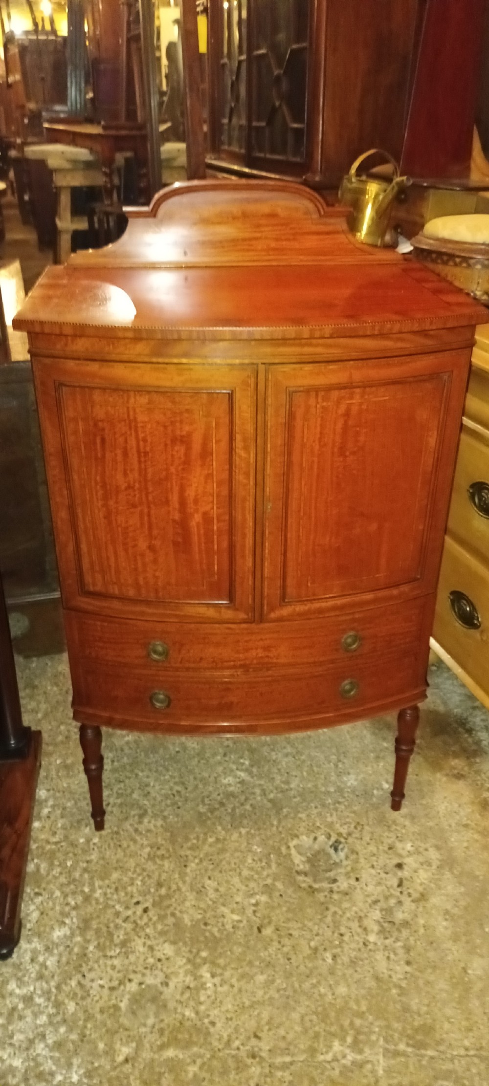 An Edwardian bowfront satinwood cabinet featuring inlaid door panels, two lower drawers, brass drop handles, and turned legs. View IV
