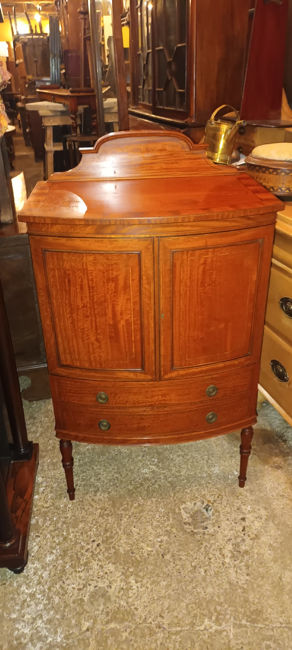 An Edwardian bowfront satinwood cabinet featuring inlaid door panels, two lower drawers, brass drop handles, and turned legs. View V
