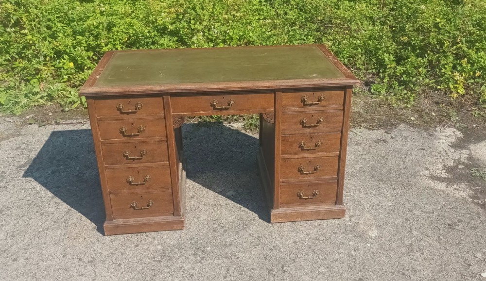 A late Victorian oak kneehole pedestal writing desk featuring nine drawers with brass handles and a green leather insert on the top surface. View X