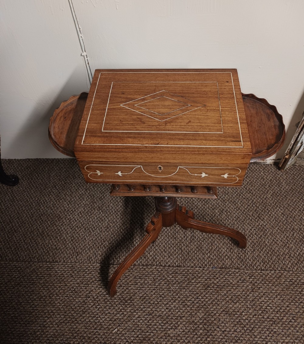 An Anglo-Indian rosewood work table featuring a hinged lid with velvet lining, fitted internal compartments, bone inlay, drop-leaf flaps, a spindle gallery, and a tripod pedestal base. View IX