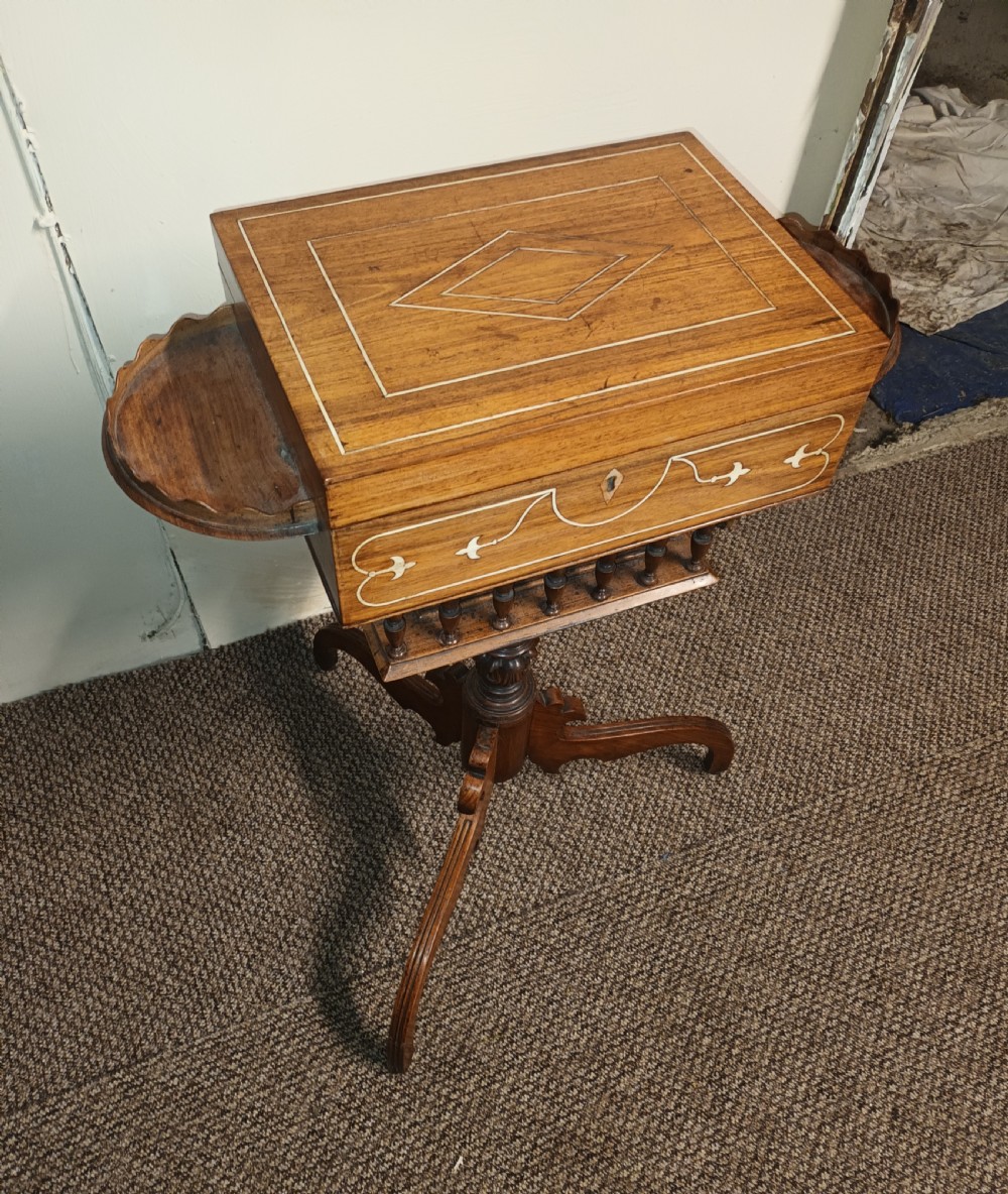 An Anglo-Indian rosewood work table featuring a hinged lid with velvet lining, fitted internal compartments, bone inlay, drop-leaf flaps, a spindle gallery, and a tripod pedestal base. View XI