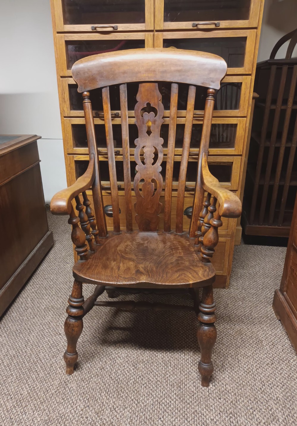 A dark-stained Victorian Windsor armchair with a spindled high back, carved central splat, shaped armrests, solid seat, and turned wooden legs. View V