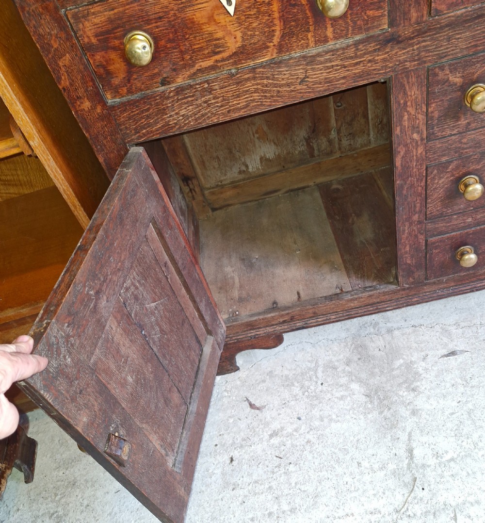 An early 19th-century dark oak Welsh dresser featuring a three-tier open shelving rack above a base with multiple drawers, brass knobs, bone escutcheons, and panelled cupboard doors. View II