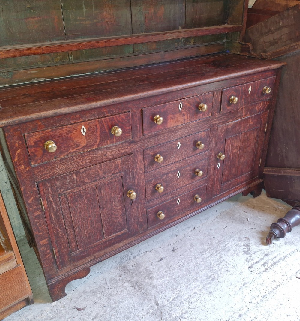 An early 19th-century dark oak Welsh dresser featuring a three-tier open shelving rack above a base with multiple drawers, brass knobs, bone escutcheons, and panelled cupboard doors. View V