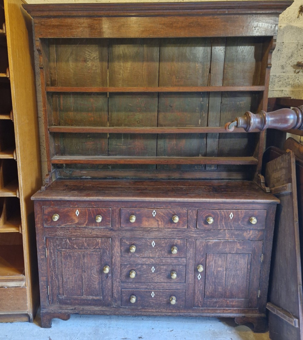 An early 19th-century dark oak Welsh dresser featuring a three-tier open shelving rack above a base with multiple drawers, brass knobs, bone escutcheons, and panelled cupboard doors. View VII