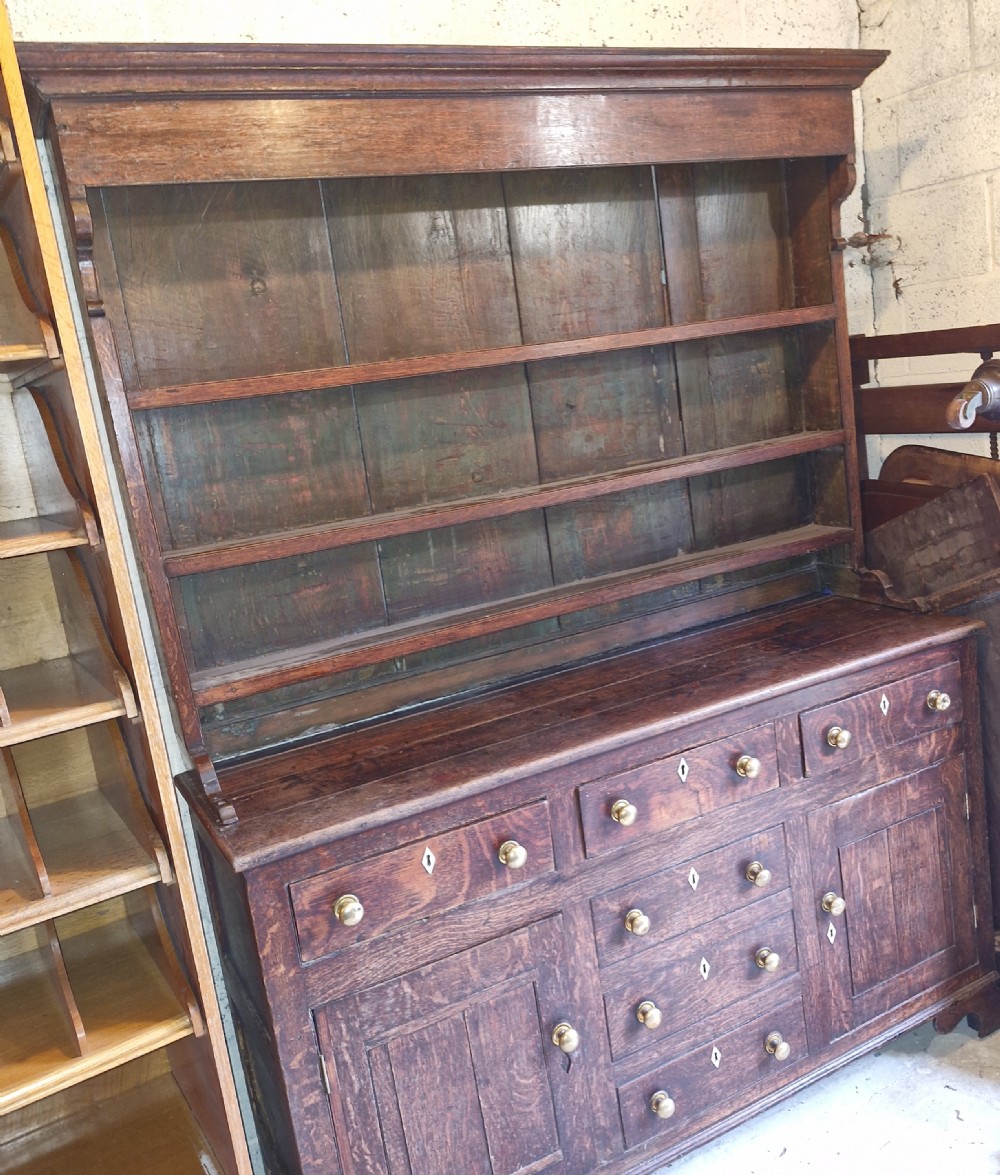 An early 19th-century dark oak Welsh dresser featuring a three-tier open shelving rack above a base with multiple drawers, brass knobs, bone escutcheons, and panelled cupboard doors. View VIII