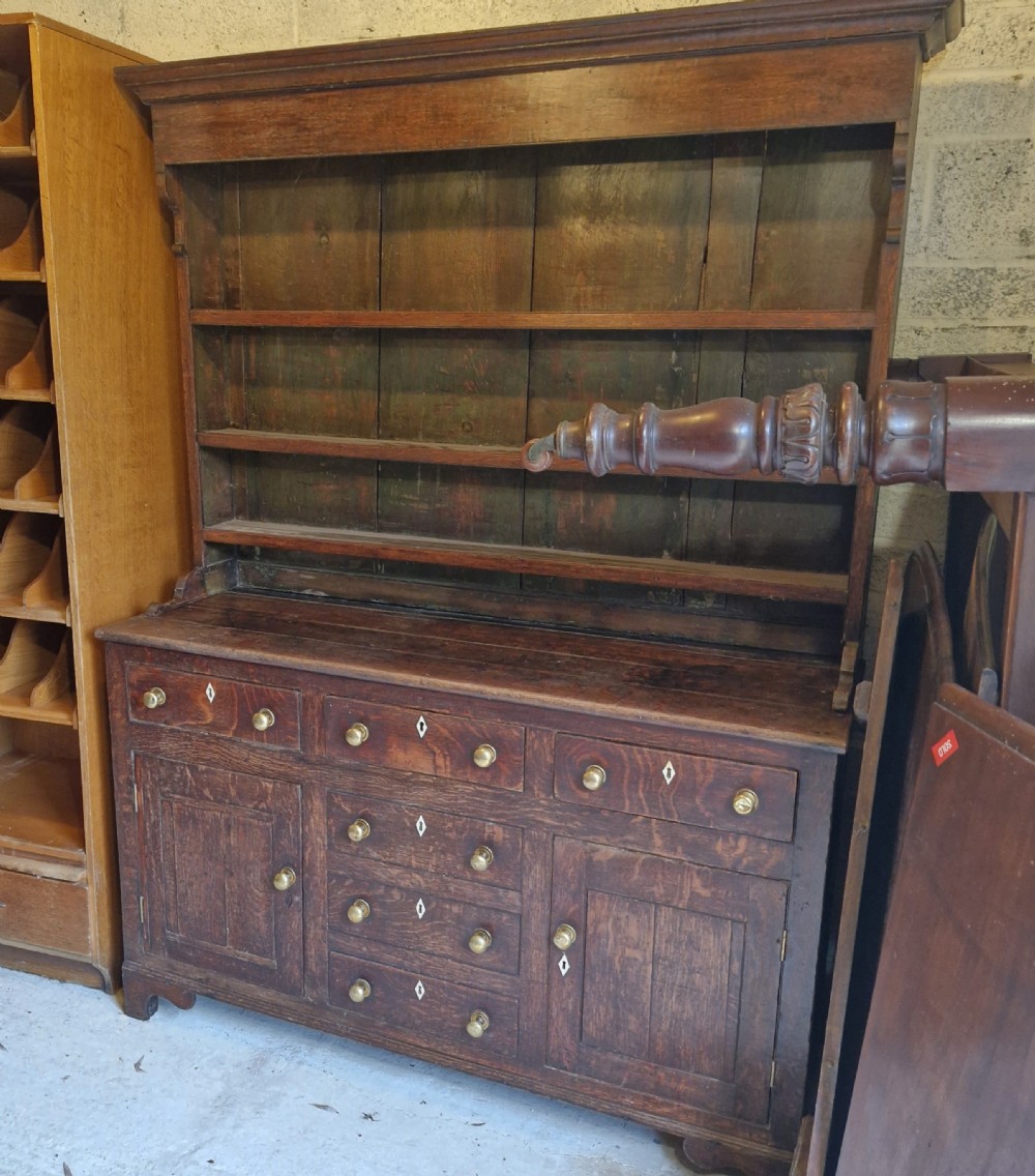 An early 19th-century dark oak Welsh dresser featuring a three-tier open shelving rack above a base with multiple drawers, brass knobs, bone escutcheons, and panelled cupboard doors. View XII