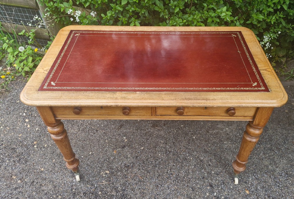 A Victorian oak writing table featuring a burgundy leather inset top, two drawers with wooden knobs, turned legs, and brass castors. View V