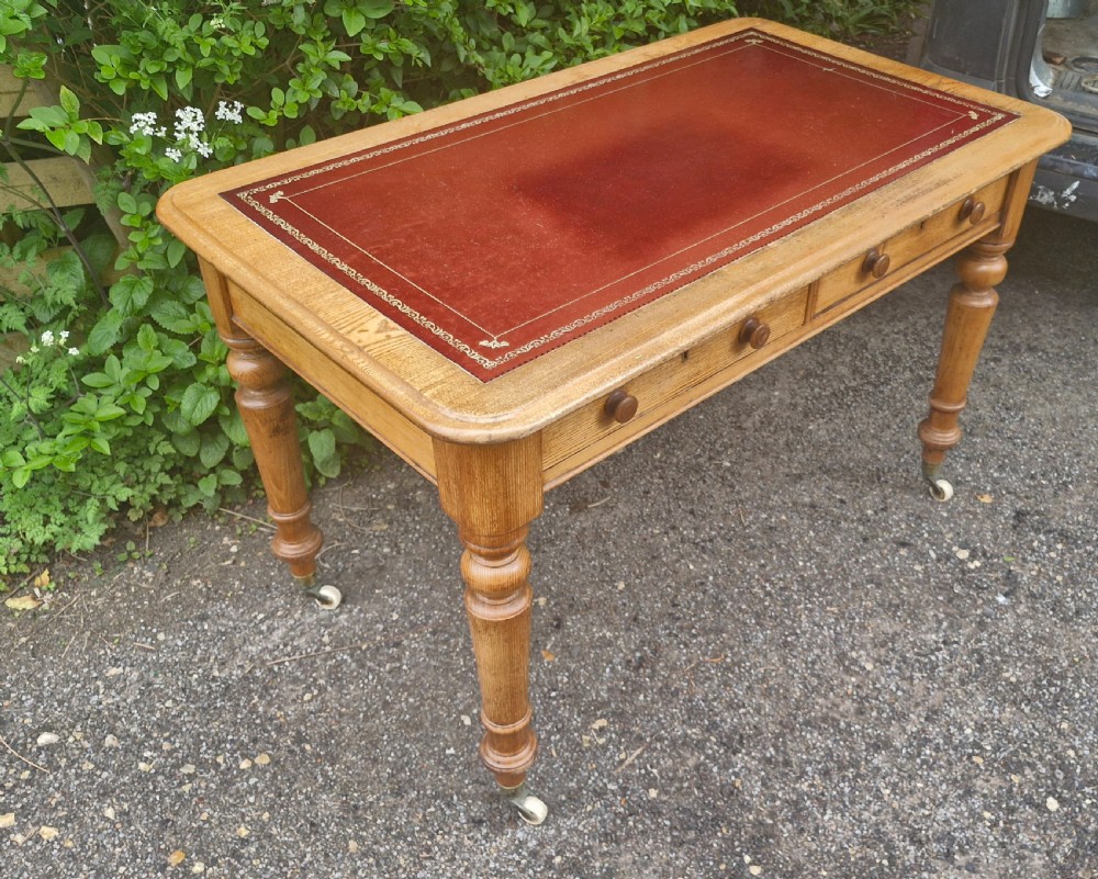 A Victorian oak writing table featuring a burgundy leather inset top, two drawers with wooden knobs, turned legs, and brass castors. View VI
