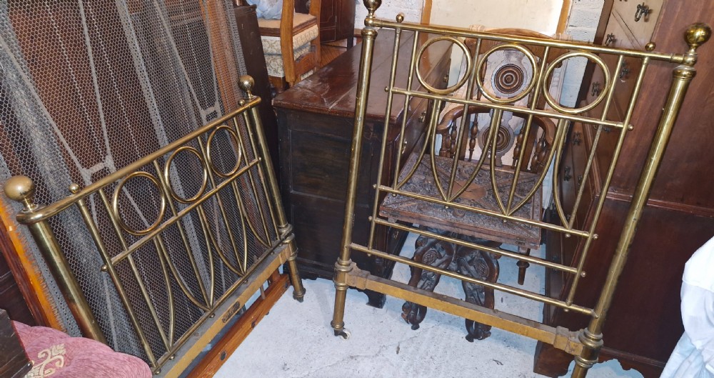 A Victorian brass single bed frame featuring geometric circular motifs, vertical spindles, brass finials, and castors. View II