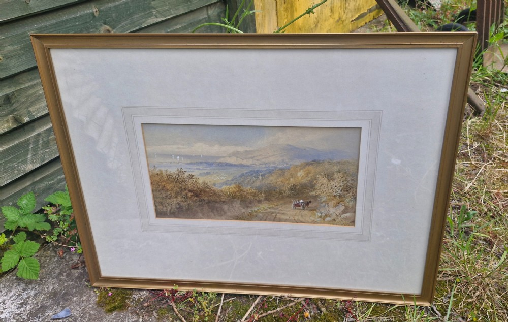 A framed 19th-century watercolour landscape painting showing a horse-drawn cart on a dirt track overlooking hills and the sea, housed within a wide mount and a slim gold frame.