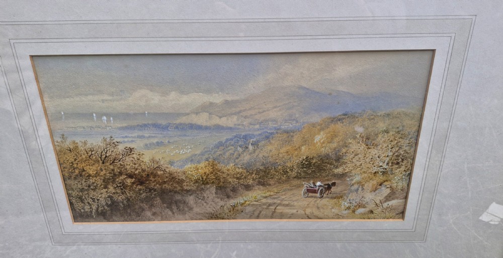 A framed 19th-century watercolour landscape painting showing a horse-drawn cart on a dirt track overlooking hills and the sea, housed within a wide mount and a slim gold frame. View II