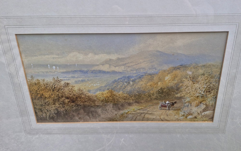 A framed 19th-century watercolour landscape painting showing a horse-drawn cart on a dirt track overlooking hills and the sea, housed within a wide mount and a slim gold frame. View VI