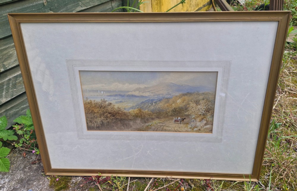 A framed 19th-century watercolour landscape painting showing a horse-drawn cart on a dirt track overlooking hills and the sea, housed within a wide mount and a slim gold frame. View VIII