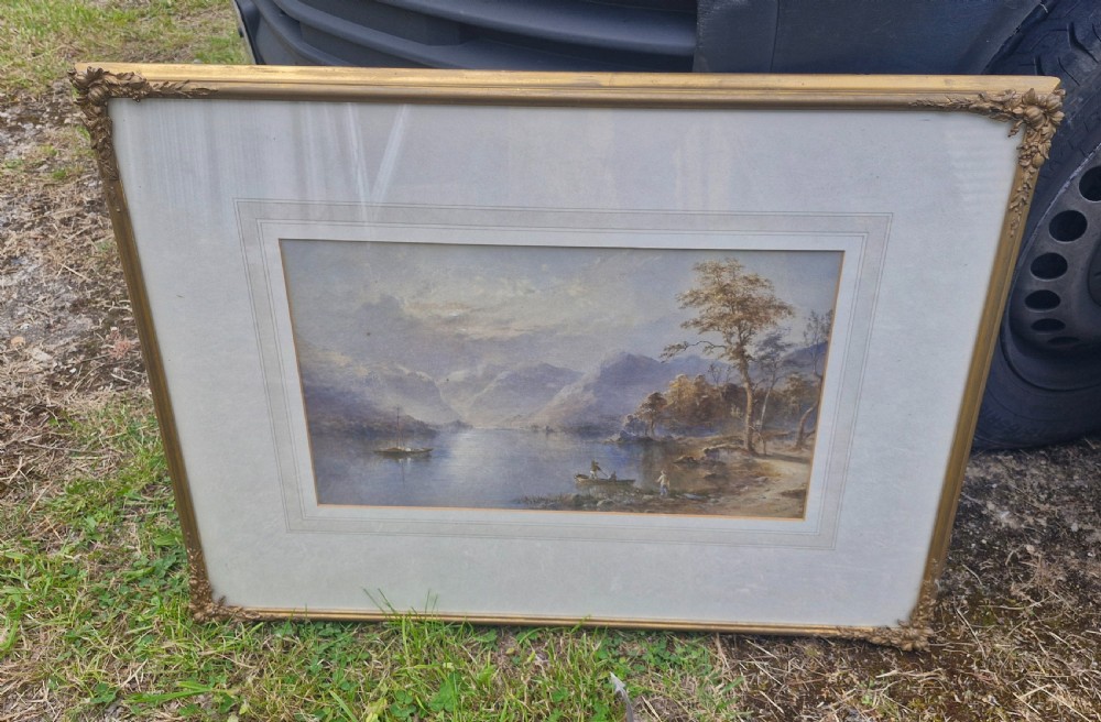 A framed 19th-century watercolour painting depicting a Lake District scene with small boats on a lake surrounded by trees and mountains, enclosed in a cream wash mount and a gilded decorative frame.