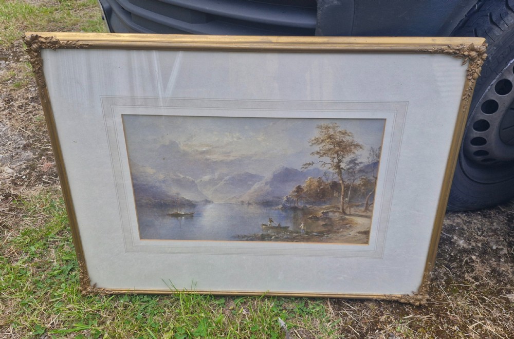 A framed 19th-century watercolour painting depicting a Lake District scene with small boats on a lake surrounded by trees and mountains, enclosed in a cream wash mount and a gilded decorative frame. View II