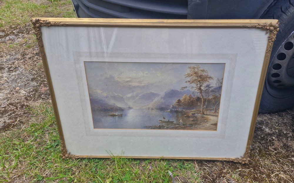 A framed 19th-century watercolour painting depicting a Lake District scene with small boats on a lake surrounded by trees and mountains, enclosed in a cream wash mount and a gilded decorative frame. View III