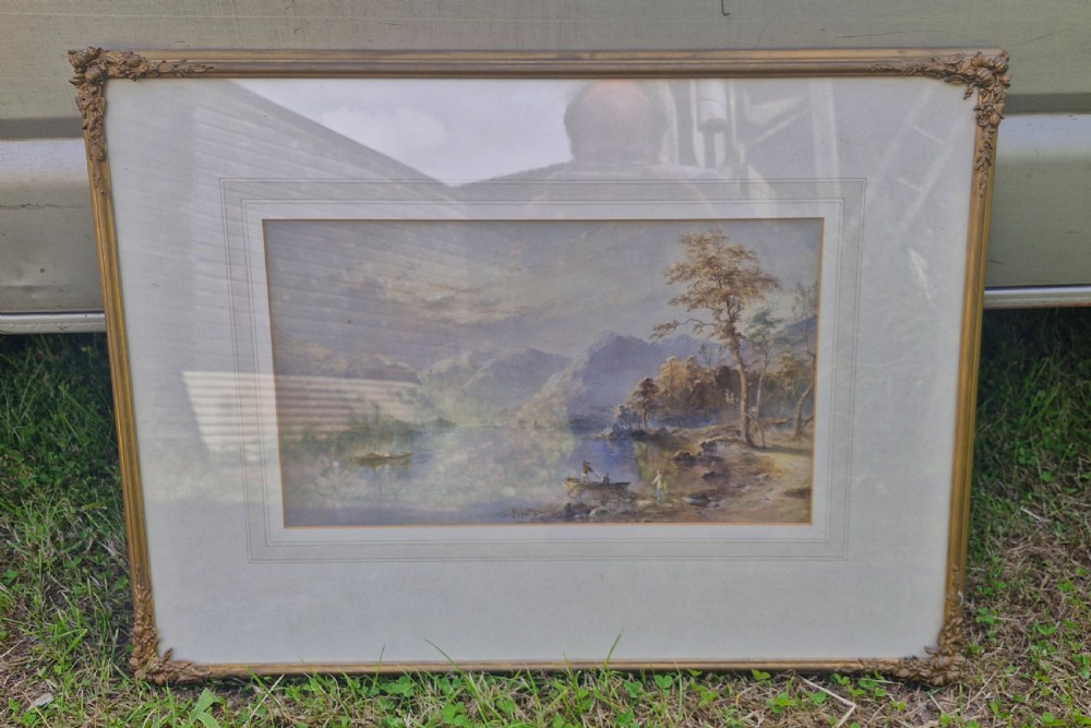 A framed 19th-century watercolour painting depicting a Lake District scene with small boats on a lake surrounded by trees and mountains, enclosed in a cream wash mount and a gilded decorative frame. View VI
