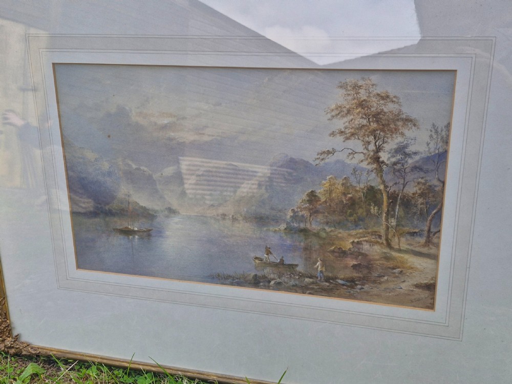 A framed 19th-century watercolour painting depicting a Lake District scene with small boats on a lake surrounded by trees and mountains, enclosed in a cream wash mount and a gilded decorative frame. View VII